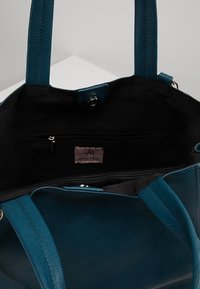 Tyrkysově modrá kabelka z texturovaného materiálu, s prostorným černým vnitřkem, který obsahuje kapsu na zip a koženou značkovou visačku.