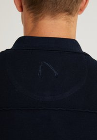 Mörkblå stickad polo med krage och synlig sömnadsdetalj på baksidan, med en liten fläck av lösa trådar.