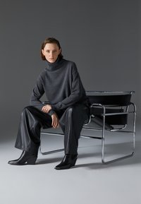 Suéter gris de cuello alto con un ajuste relajado, combinado con pantalones de cuero negro y botas de cuña, sentado en una silla con marco de cromo.