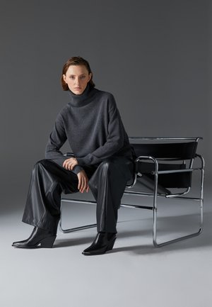 Pullover grigio con colletto a dolcevita e vestibilità comoda, abbinato a pantaloni in pelle nera e stivali con zeppa, seduto su una sedia con struttura in cromo.