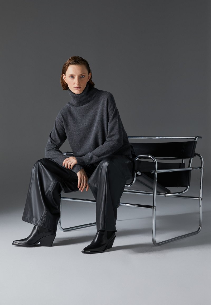 Suéter gris de cuello alto con un ajuste relajado, combinado con pantalones de cuero negro y botas de cuña, sentado en una silla con marco de cromo.