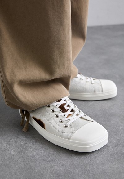 White sneakers made of textured material, featuring brown patches, round toe, and white rubber sole; paired with tan, loose-fitting pants.
