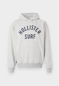 Svijetlosivi dukserica s kapuljačom i prednjim džepom, s tamnoplavim natpisom "Hollister Surf" preko prsa.