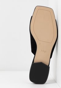 Semelle d'une chaussure plate noire reposant contre un mur blanc, montrant une semelle beige lisse, un talon noir texturé et la marque "högI".