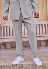 Lichtbeige op maat gemaakte kostuum met een dubbelgeknopte blazer en bijpassende broek. Draagt een combinatie met witte sneakers, een gladde textuur en een rechte pasvorm.