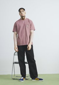 T-shirt rose à manches courtes avec une poche, associé à un pantalon noir et des baskets bleues avec des rayures jaunes. Le modèle se tient devant un fond blanc.