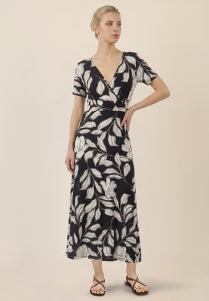 Frau in schwarzem Wickelkleid mit großem weißem Blumenmuster, kurzen Ärmeln und knöchellangem Saum, trägt braune Riemchensandalen, steht mit der Hand in der Hüfte.