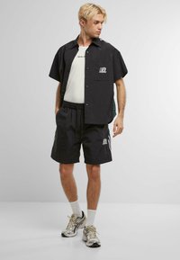 Sort kortærmet skjorte med knaplukning og grønne detaljer, parret med sorte shorts. Har et logo og elastisk talje. Sølv sneakers fuldender looket.