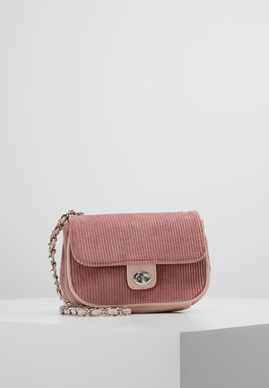 Mała lawendowa torba na ramię z aksamitu prążkowanego z klapką, srebrną zapinką na przekręcanie i łańcuszkiem, leżąca na białej powierzchni na jednolitym tle.