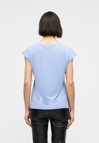 Γυναίκα με κοντά σκούρα μαλλιά, που φοράει ανοιχτό γαλάζιο μπλουζάκι με κοντά μανίκια και μαύρο δερμάτιν ο παντελόνι, με την πλάτη στραμμένη σε απλό φόντο.