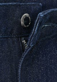 Tela de mezclilla de color azul oscuro con una textura suave, que presenta un botón de metal plateado y un detalle de cremallera en el frente.