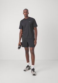 Schwarzes Nike T-Shirt und Shorts, athletic Fit. Graue Sneaker mit schwarzen Akzenten und weißen Socken. Hält eine schwarze Wasserflasche. Weißer Hintergrund.