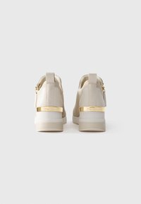 Un paio di sneaker alte beige con tessuto a maglia, dettagli dorati sul tallone, cerniere e anelli sul tallone con marchio, viste dal retro.