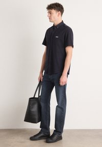 Camicetta blu navy a maniche corte con logo "BOSS", abbinata a jeans scuri e sneakers nere, mentre tiene una borsa tote nera.