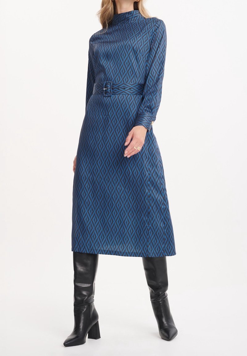 Niebieska sukienka w wzory z długimi rękawami, z wysoką szyją, paskiem w talii i diamentowymi wzorami; w połączeniu z czarnymi botkami na obcasie.