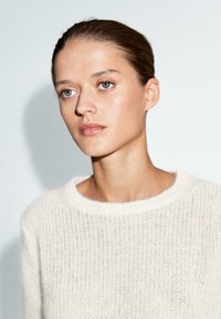 Maglione lavorato a maglia beige chiaro con scollatura rotonda e una texture a maglia larga, caratterizzato da un aspetto morbido e arioso. Il modello ha la pelle liscia e capelli chiari.