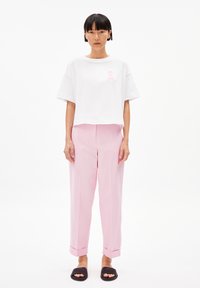 Witte crop-top met een roze graphic op de borst, gecombineerd met lichtroze geplooide broeken en zwarte slipper sandalen. Zachte stoftexturen.