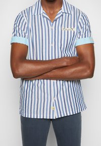 Camisa de manga corta con botones en rayas verticales azules y blancas, con puño azul claro, bolsillo en el pecho y un logo de sol en el dobladillo.