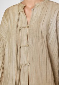 Blouse beige texturée avec une fermeture à moitié boutonnée et des liens décoratifs. Tissu doux et léger avec des plis subtils et une coupe décontractée.