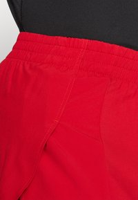 Rote Shorts mit einem elastischen Bund, aus glattem, leichtem Stoff und mit kontrastierenden gelben Nähten an den Säumen.