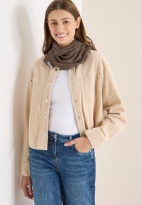 Veste-chemise en velours côtelé beige avec boutons-pression, deux poches, portée sur un haut blanc et associée à un jean bleu ; écharpe marron drapée autour du cou.