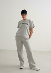 Grå sportstøjsæt bestående af en kortærmet t-shirt med teksten "Sportswear" og brede bukser, parret med en sort kasket og hvide sneakers.