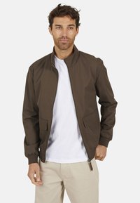 Veste zippée marron avec col montant, deux poches avant, poignets élastiques et texture lisse. Portée sur un t-shirt blanc et un pantalon beige.