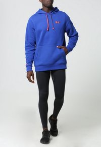 Niebieska bluza z kapturem i pomarańczowymi sznurkami, z kieszenią z przodu, ściągaczami przy rękawach, zestawiona z czarnymi legginsami i czarnymi sneakersami.