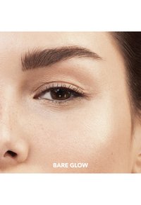 Nærbilde av et øye med definerte øyenbryn, lysebrune vipper og en glatt, fuktig hudtekstur. Teksten under leser "BARE GLOW."