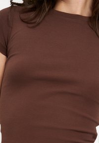 Gros plan d'une personne portant une chemise marron cintrée à manches courtes montrant une partie du cou et du haut du torse avec des cheveux foncés visibles.