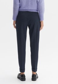 Osoba viděná zezadu, oblečená v tmavých strukturovaných kalhotách s manžetami, levandulovém svetru a černých mokasínech na bílém pozadí.