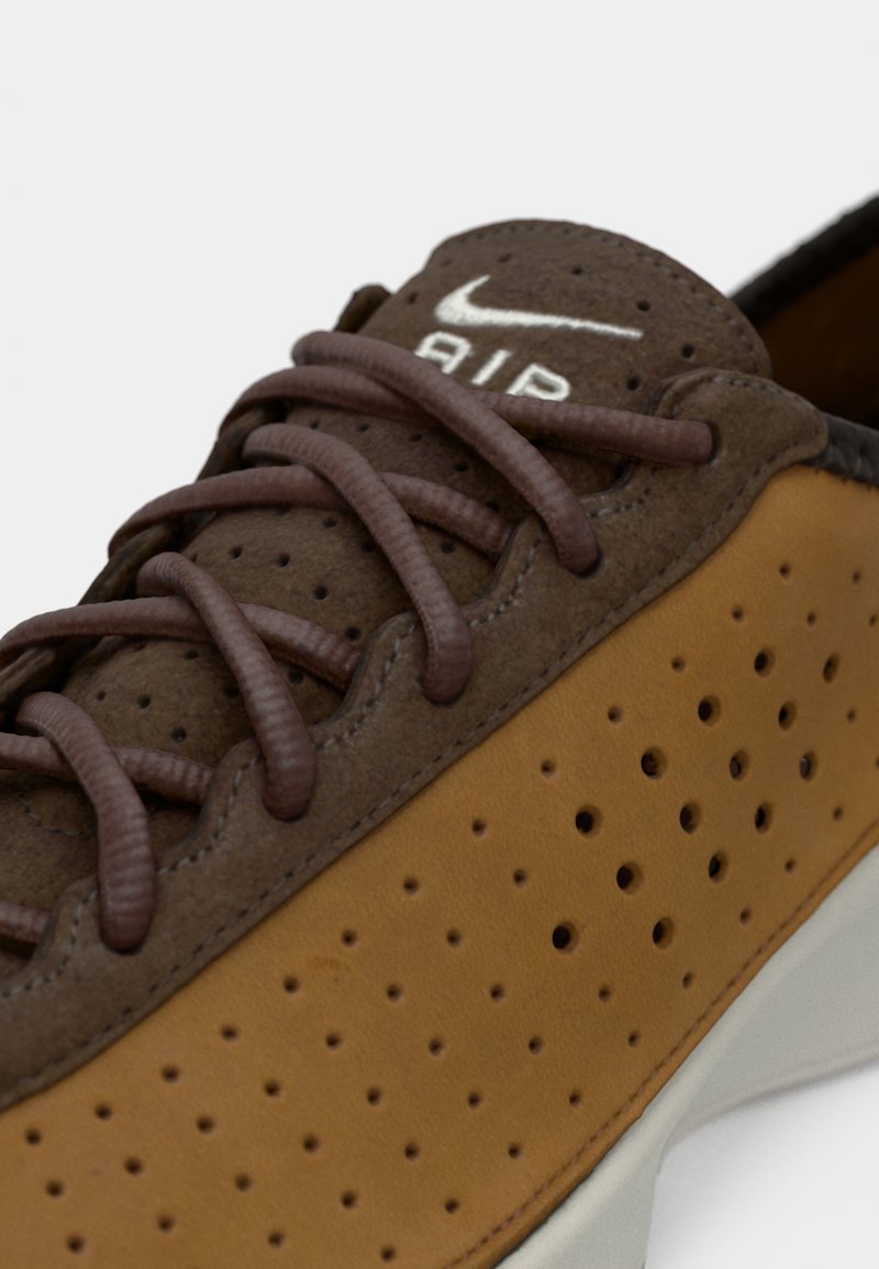 Des chaussures Nike avec une tige en daim marron, des panneaux perforés beiges, des lacets marron et un logo blanc, surmonté d'une semelle de couleur claire.