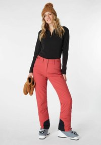 Pantaloni da sci rossi con un design aderente, abbinati a un top nero a maniche lunghe e stivali da sci grigi. Gli accessori includono un berretto in maglia marrone e guanti.