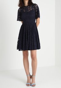 Marinblå spetsklänning med korta ärmar, figurnära överdel och utsvängd kjol. Har blommönster och en delikat textur. Bärs med ljusblå klackar.