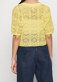 Gul stickad topp med korta puffärmar, med ett öppet, spetsliknande mönster. Matchad med mörkblå högmidjade jeans.