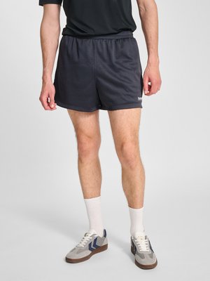 Man die zwarte sportshorts, witte sportsokken, grijze en marineblauwe sneakers en een zwart T-shirt draagt, staande tegen een effen achtergrond.