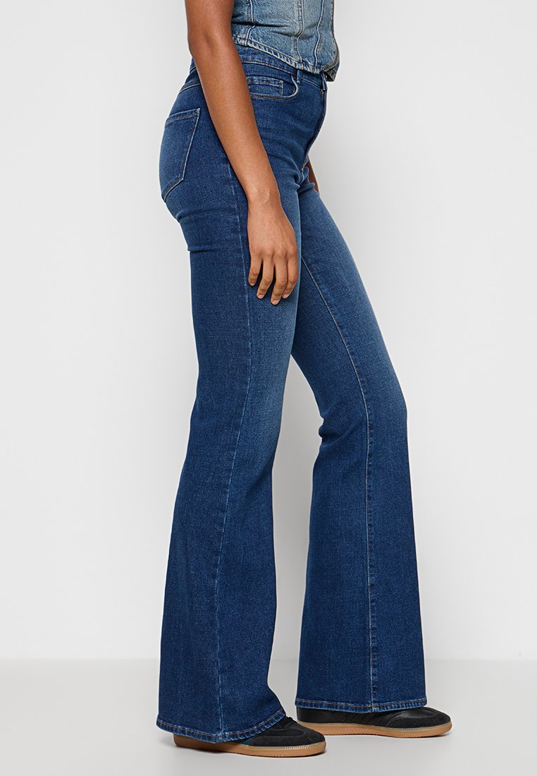 Femme portant un jean en denim bleu foncé ajusté avec des jambes évasées et des chaussures noires, se tenant de profil devant un fond uni.