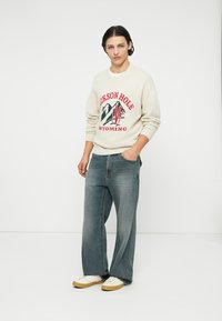 Beżowy sweter z dzianiny z czerwono-zielonym napisem "Jackson Hole Wyoming", zestawiony z luźnymi ciemnymi dżinsami i białymi trampkami.