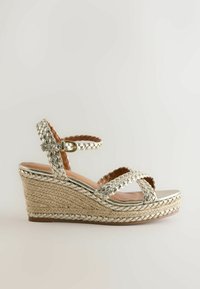 Sandale compensée avec brides métalliques tressées et boucle à la cheville, talon plateforme enveloppé de jute, semelle intérieure beige, vue de côté sur fond uni.