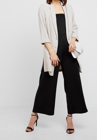 Blazer beige clair à manches longues, ouvert sur le devant avec des revers, porté sur une combinaison noire cintrée à jambes larges. Talons noirs à brides et pochette blanche.