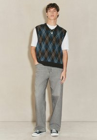Mehrfarbiger Argyle-Weste in Schwarz, Blau und Braun über einem weißen T-Shirt, kombiniert mit grauen weitgeschnittenen Jeans und schwarz-weißen Turnschuhen.