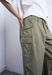 Άτομο που φοράει ελαιοπράσινο παντελόνι cargo με τσέπες με καπάκι και λευκό πουκάμισο φορεμένο μέσα, με το χέρι να ακουμπά στην πίσω τσέπη, στέκεται σε εσωτερικό χώρο.