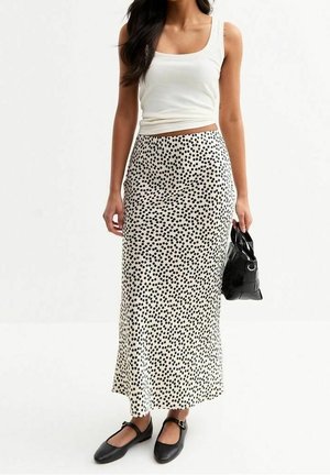 Donna con top bianco senza maniche, gonna lunga nera a pois bianchi, ballerine nere e che tiene una piccola borsa nera.