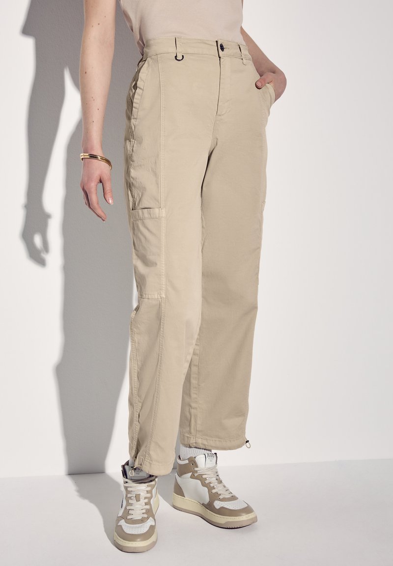 Beige Cargo-Hosen mit zwei seitlichen Taschen, Reißverschluss-Bündchen, hohem Taillenbund und lockerer Passform, kombiniert mit beige-weißen High-Top Sneakers.