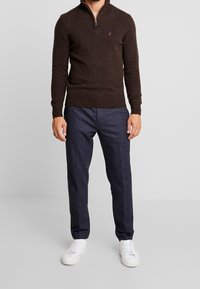 Brun zip-up tröja med ribbade muddar och nederkant, i kombination med mörka strukturerade byxor och vita sneakers. Tröjan har en liten logodetalj.