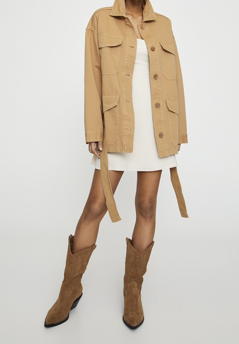 Femme portant une veste utilitaire beige sur une robe blanche, associée à des bottes de cowboy en daim marron, debout devant un fond clair uni.