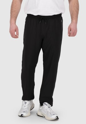 Schwarze Sporthosen mit einem elastischen Bund und Kordelzug, ausgestattet mit Reißverschlusstaschen und einer glatten Textur. Getragen mit weißen Sneakers.