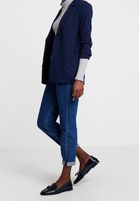 Blazer bleu marine à coupe ample, deux poches avant, associé à un col roulé gris clair, jean bleu retroussé, et mocassins noirs brillants.