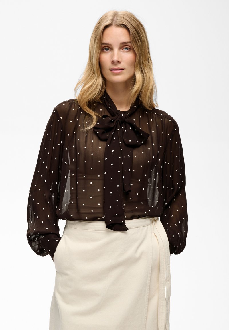 Femme blonde portant un chemisier noir transparent à pois avec un grand nœud papillon et une jupe crème taille haute avec des poches.