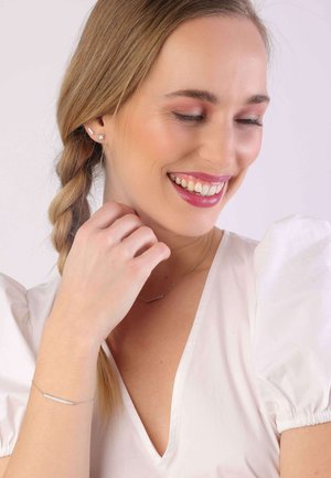 Bijoux en argent comprenant un bracelet fin et droit et un collier délicat avec une barrette horizontale. Le mannequin porte un haut blanc à manches bouffantes.